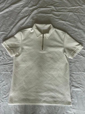 Zip tröja - Vit topp med korta ärmar och halv dragkedja framtill. Toppen har ett upphöjt geometriskt strukturmönster och stårkrage. Materialet känns mjukt och stretchigt, perfekt för en clean och modern look.