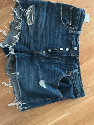 Levi's denimshorts  - Säljer ett par blå denimshorts från Levi's med hög midja och slitna detaljer. Shortsen har råa kanter, knappgylf och klassiska  fickor. Perfekta för sommaren och ger en avslappnad vibe. W:31. 