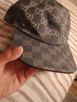 Blå keps - Säljer en snygg keps från Gucc* i mörkgrått tyg med det klassiska -monogrammet över hela kepsen. Kepsen har böjd skärm och justerbar passform. Materialet är slitstarkt canvas och insidan är fodrad i ljus textil. Perfekt för dig som gillar exklusiva accessoarer.