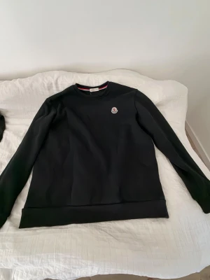 Svart sweatshirt från Moncler - Svart sweatshirt från Moncler med rund halsringning och diskret logotyp på bröstet. Tröjan har ribbade muddar vid ärmslut och nederkant samt en klassisk passform. Perfekt för dig som gillar stilrena och exklusiva plagg.