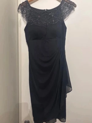 Svart klänning med pärlor DJ-Jaz - Supersnygg svart klänning från DJ-Jaz med pärlbroderad mesh upptill och elegant drapering på sidan. Klänningen har en tight passform och är perfekt för dig som vill sticka ut med detaljer som pärlor och transparent mesh vid axlarna.
