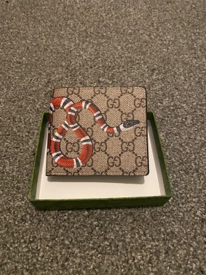  plånbok med orm och GG-mönster - Snygg plånbok i beige canvas med klassiskt GG-mönster och en cool röd, vit och svart orm på framsidan. Insidan är mörkbrun med flera kortfack och ett fönster för ID-kort. Perfekt för dig som gillar lyxiga accessoarer med unik design.