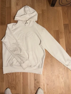 Vit hoodie från Polo Ralph Lauren - Snygg vit hoodie från Polo Ralph Lauren med broderad logga på bröstet. Klassisk känguruficka framtill, ribbade muddar och huva med dragsko. Perfekt för en avslappnad och stilren look. Mjuk bomullsblandning som känns skön mot huden.
