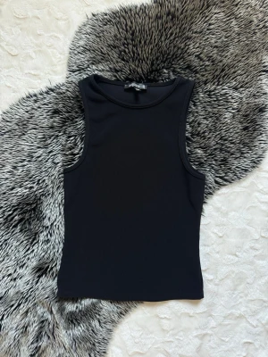 Svart ribbad linnetopp från Gina Tricot - En stilren svart linnetopp med ribbad struktur. Toppen är ärmlös och har en tight passform som framhäver figuren. Perfekt att matcha med jeans eller kjol för en clean look.