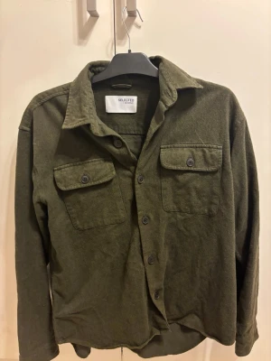Olivgrön overshirt från Selected Homme - Stilren olivgrön overshirt från Selected Homme med två bröstfickor med lock och knappar. Skjortjackan har klassisk krage, raka ärmar och knäppning framtill. Tillverkad i mjukt tyg som ger en avslappnad look.