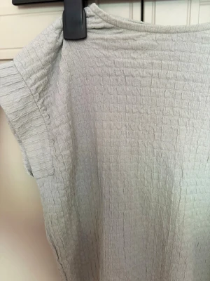 Ljusgrå topp med struktur från Zara - En ljusgrå topp från H&M, har gröna drag i sig, med korta ärmar och en snygg strukturerad yta. Toppen har en avslappnad passform och är tillverkad i ett mjukt material som känns skönt mot huden. Perfekt att styla med jeans eller kjol. Baksidan är lite öppen med fina detaljer-rosetter.