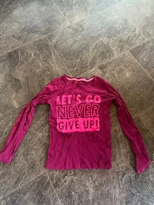 Rosa långärmad topp med tryck - Cool långärmad topp från H&M i en stark rosa färg med texten 'LET'S GO NEVER GIVE UP!' tryckt framtill i olika nyanser av rosa. Rund halsringning och mjukt bomullsmaterial som är skönt att bära. Perfekt för dig som gillar peppiga budskap och färg! (Plus frakt) 