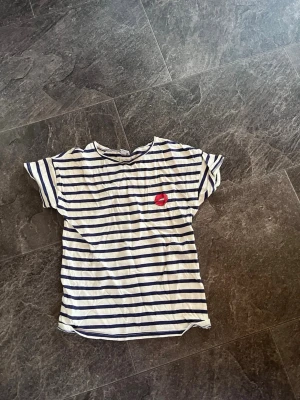Randig t-shirt med läppmotiv - Vit och marinblå randig t-shirt med korta ärmar och ett rött läppmotiv på bröstet. Avslappnad passform och rund halsringning, perfekt för en chill vardagslook. (Plus frakt) 