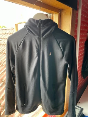 Svart hoodie från Peak Performance - Snygg svart hoodie från Peak Performance med dragkedja framtill och två fickor med dragkedja. Liten logga på bröstet och texten 'PERFORMANCE' på huvan. Tillverkad i mjukt och stretchigt material, perfekt för en sportig och avslappnad stil.
