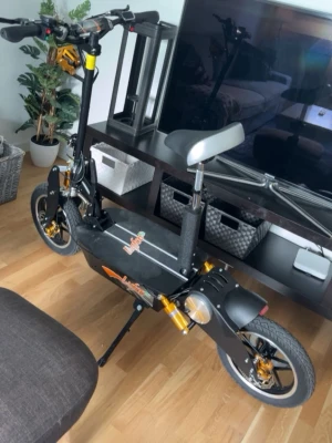 Svart el-scooter med sadel - Elektrisk scooter i svart med sadel och gulddetaljer. Scootern har breda däck, fotplatta och styre med handtag. Märket Joyor syns på sidan. Perfekt för smidig transport i stan.