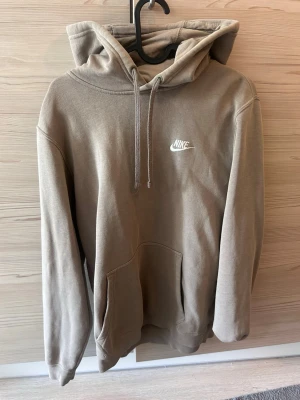Beige hoodie från Nike med huva - Snygg beige hoodie från Nike med klassisk logga broderad i vitt på bröstet. Hoodien har stor huva med dragsko och en rymlig magficka. Tillverkad i mjukt bomullsmaterial och har långa ärmar med ribbade muddar. Perfekt för en avslappnad och trendig stil.