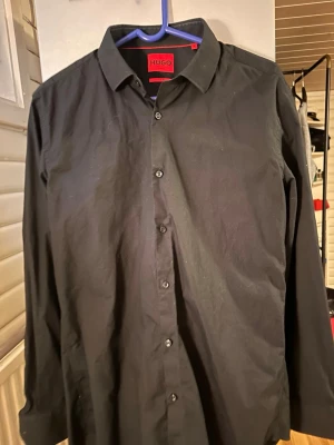 Svart skjorta från HUGO - Klassisk svart skjorta från HUGO med normal passform och knappar framtill. Skjortan har lång ärm och stilren design, perfekt för dig som gillar en clean och enkel look. Tillverkad i bomull för en bekväm känsla. Storlek M/38