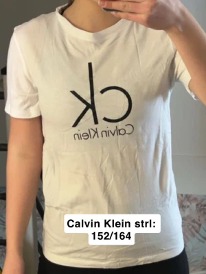 Vit Calvin Klein t-shirt med logga - Säljer en vit t-shirt från Calvin Klein i storlek 152/164. T-shirten har klassisk passform, korta ärmar och stor svart CK-logga framtill. Tillverkad i mjuk bomull som känns skön mot huden.  30 kr varav en lite gul fläck som jag är osäker på om den går bort 