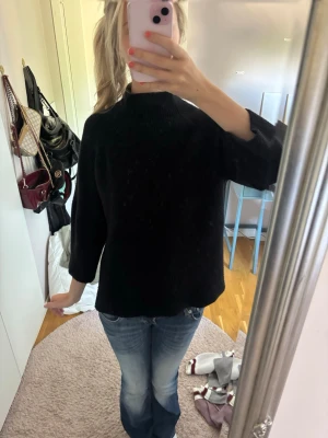 Svart stickad tröja med halvpolokrage - En svart stickad tröja med halvpolokrage och trekvartsärmar. Tröjan har en rak passform och är enkel men stilren, perfekt att matcha med jeans eller kjol. Materialet ser mjukt och bekvämt ut, och färgen är helsvart utan mönster.