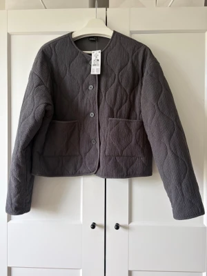 Grå quiltad cardiganjacka från Gina Tricot - Snygg grå quiltad cardiganjacka från Gina Tricot i färgen Asphalt. Jackan har rund halsringning, knäppning framtill med fyra knappar och två stora fickor. Materialet är strukturerat och ger en modern look. Perfekt för lager-på-lager-stil.