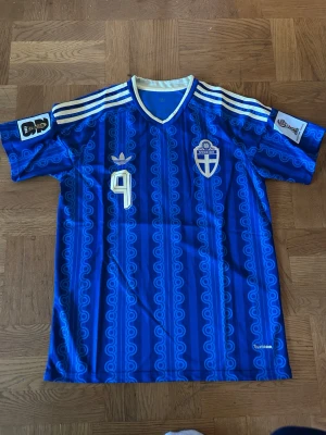 Blå Adidas fotbollströja Grekland - Snygg blå fotbollströja från Adidas med Greklands landslagsemblem, nummer 9 på bröstet och vita detaljer. Tröjan har klassiska Adidas-ränder på axlarna och ett unikt mönster över hela tyget. Perfekt för fotboll eller streetwear.