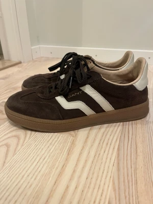 Bruna Gant sneakers med vita detaljer - Snygga bruna sneakers från Gant i mocka med vita läderdetaljer och klassiska ränder på sidan. Skorna har bruna snören och en gummisula i naturfärg. Perfekta för en stilren och avslappnad look.