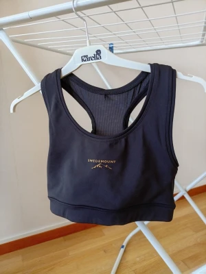 Svart sport-bh från Swedemount - Sport-bh i svart från Swedemount med brottarrygg och meshdetaljer för extra ventilation. Bh:n har en stilren design med logga framtill och bred resår nedtill för bra stöd. Perfekt för träning och aktiva dagar.