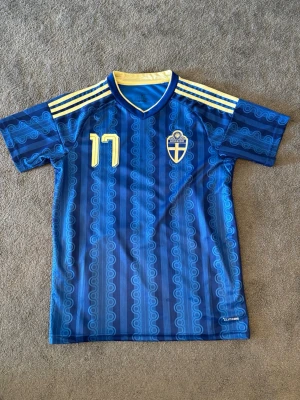 Blå Sverige fotbollströja Adidas - Säljer en blå Sverige fotbollströja från Adidas med nummer 17 och namnet Gyökeres på ryggen. Tröjan har gula detaljer, mönstrad design och tre gula ränder på axlarna. Perfekt för dig som vill stötta landslaget med stil. Som ny. Storlek m. 