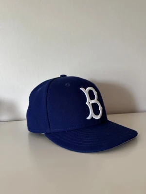 Blå keps med vit B-logga - Hej säljer min snygg blå keps med lite böjd skärm och stor vit broderad B-logga framtill (Boston). Klassisk basebollmodell som passar perfekt till streetwear eller sportiga outfits. Den är köpt i USA och är i bra skick. Hör av er vid frågor eller funderingar :)