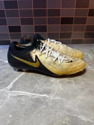 Nike phantom Elite  fotbollsskor svart/guld - Säljer ett par Nike Mercurial fotbollsskor i svart och guld med vita snören. Skorna har en låg profil, stickad ovandel och tydlig Nike-logga i guld på sidan. Perfekta för snabba spelare som vill ha bra grepp och stil på planen.