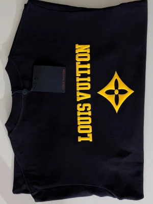 Iouis Vuitton T-shirt  - Mörkblå t-shirt i bomull med gult tryck. Framsidan pryds av en centrerad logotyp, och baksidan har ett större texttryck tillsammans med en grafisk blomstermodul. Tröjan har en klassisk passform med rund halsringning och korta ärmar.( Fast pris)