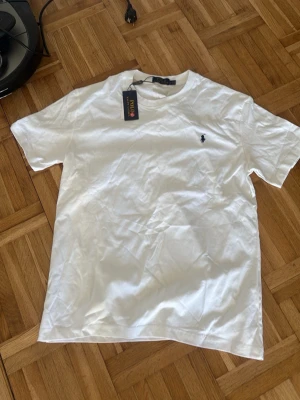 Vit t-shirt från Polo Ralph Lauren - Klassisk vit t-shirt från Polo Ralph Lauren med den ikoniska lilla loggan broderad på bröstet. T-shirten har rund hals, korta ärmar och är gjord i mjuk bomull. Perfekt basplagg för en clean och stilren look.