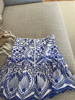 Vit och blå kjol från Zara XS - Supersöt kort kjol från Zara i vitt och blått med dekorativa knappar framtill och volangkant. Kjolen har ett unikt mönster och är perfekt för sommarens alla tillfällen. Tillverkad i ett lätt och skönt material.
