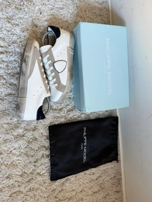 Philippe Model vita och beige sneakers - Snygga sneakers från Philippe Model i vitt och beige läder med mörkblå detaljer på hälen. Klassisk låg modell med vita snören och gummisula. Kommer med originalkartong och dustbag. Perfekta för dig som gillar stilrena och trendiga skor.