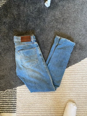 Blå slim fit jeans med läderpatch - Säljer ett par klassiska blå slim fit jeans med fem fickor och en brun läderpatch bak i midjan. Jeansen har en något tvättad look och raka ben. Perfekta för en avslappnad och trendig stil.