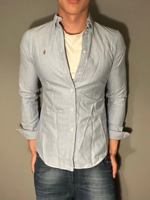 Ljusblå skjorta från Polo Ralph Lauren - Säljer en ljusblå skjorta från Polo Ralph Lauren med klassisk knappstängning och broderad logga på bröstet. Skjortan har långärmad design och är tillverkad i bomull. Perfekt för dig som gillar stilrena plagg med snygga detaljer.
