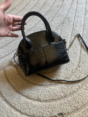 Svart croco-mönstrad handväska - Cool svart handväska med croco-mönster och glänsande finish. Väskan har både korta handtag och en lång axelrem med kedjedetaljer. Perfekt för dig som gillar stilrena accessoarer med lite edge. Materialet är konstläder. Finns ej att köpa längre🩷