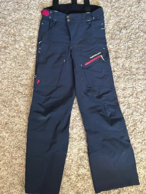 Blå täckbyxor från Peak Performance - Snygga mörkblå skalbyxor från Peak Performance med cerisa detaljer och dragkedjor. Byxorna har hängslen, flera fickor med dragkedja och förstärkta knän. Perfekta för skidåkning eller vinteraktiviteter. Materialet är slitstarkt och vattenavvisande. Tecken på slitage längst ned på benet där byxor går över och mot pjäxorna.