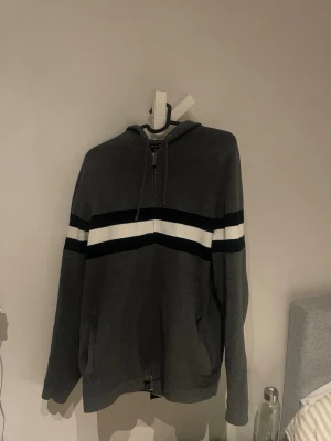 Massimo dutti zip hoodie - Mörkgrå hoodie med dragkedja framtill och huva. Två breda ränder i svart och vitt går tvärs över bröstet och armarna. Hoodien har fickor på sidorna och en avslappnad passform. Perfekt för chill dagar eller när du vill ha en enkel streetstyle-look.