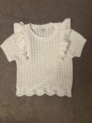 Vit stickad topp med volang från Lindex - Supersöt vit stickad topp från Lindex med korta ärmar och volangdetaljer på axlarna. Tröjan har ett fint hålmönster och vågig kant nertill och vid halsen. Storlek 146-152 