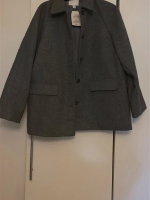Grå ullkappa från H&M - Stilren grå kappa från H&M i ullblandning. Rak passform med klassisk krage, knäppning framtill och två stora fickor. Helt ny, aldrig använd. 