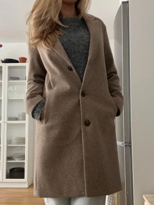 Beige kappa med knappar - Stilren beige kappa med tre knappar framtill och klassisk krage. Rak passform och diskreta fickor på sidorna. Perfekt för kyliga dagar och enkel att matcha med olika outfits. Sitter bra i storleken och är nästintill oanvänd, säljs pga används inte.