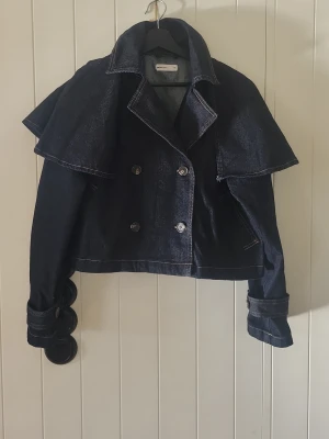 Kort denimkappa från Gina tricot  - Säljer en unik och trendig kort kappa i mörkblå denim från Gina tricot. Kappan har dubbelknäppning, bred krage och cape-liknande axelparti. Snygga detaljer med stora knappar på ärmsluten och kontrastsömmar. Perfekt för dig som vill sticka ut. Jackan är helt ny och aldrig använd. 