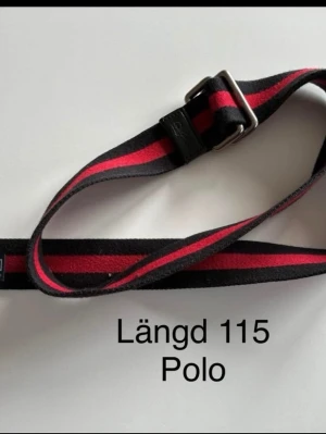 Randigt bälte från Polo, längd 115 cm - Snyggt bälte från Polo med längd 115 cm. Bältet har ett randigt mönster i svart och rött samt ett metallspänne. Perfekt accessoar för att lyfta din outfit med en sportig touch.