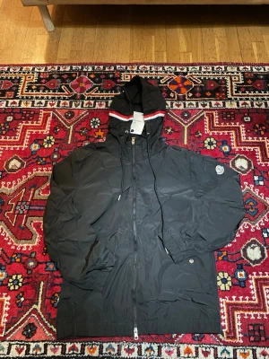 Svart vindjacka från Moncler - Snygg svart vindjacka från Moncler med huva och dragkedja framtill. Jackan har Moncler-logga på ärmen och en cool detalj med röd och vit rand på huvan. Tillverkad i lätt och tåligt material, perfekt för blåsiga dagar.