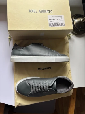 Gråa sneakers från Axel Arigato - Snygga gråa sneakers från Axel Arigato med minimalistisk design och vita sulor. Skorna är tillverkade i slätt läder och har gråa skosnören samt diskret logotyp på sidan. Perfekt för dig som gillar stilrena och moderna skor.  Låda och påse till skor ingår.