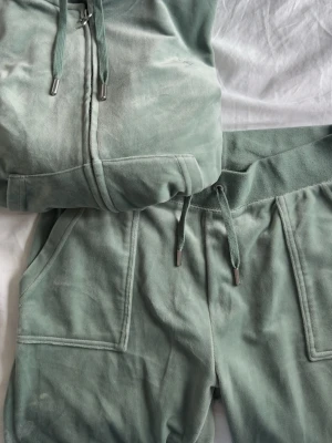 Grönt velour set Juicy Couture - Mysigt loungewear-set från Juicy Couture i ljusgrön velour. Setet består av en zip-hoodie med dragsko och matchande byxor med elastisk midja och fickor. Perfekt för chill dagar hemma. Metalländar på snören för extra detalj.