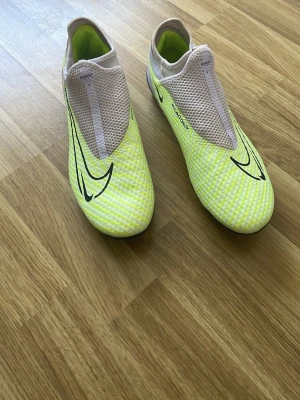 Nike Phantom GT2 fotbollsskor gul - Säljer ett par Nike Phantom GT2 fotbollsskor i en cool gul färg med svarta detaljer och mesh-paneler. Skorna har en strömlinjeformad design utan snörning och är tillverkade i syntetmaterial för optimal bollkontroll. Perfekta för spel på gräs med dobbar under sulan.