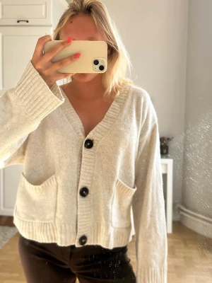 Beige kofta med stora knappar💖💖💖 - Säljer en beige, mysig kofta med tre stora svarta knappar framtill och två fickor. Koftan har breda ribbade muddar på ärmar och nederkant, samt en avslappnad passform och v-ringad hals. Perfekt för lager-på-lager och chill dagar.