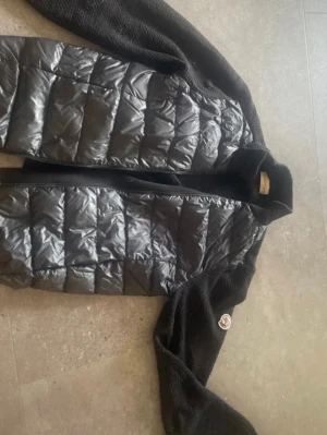 Svart dunväst från Moncler - Cool svart cardigan från Moncler med quiltad framsida i glansigt material och stickade ärmar. Västen har dragkedja med Moncler-logga och ett Moncler-emblem på ärmen. Perfekt för att höja din outfit med exklusiv känsla. Barnstorlek.