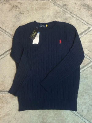Mörkblå kabelstickad tröja Polo Ralph Lauren - Polo Ralph Lauren Flätstickad Tröja - Marinblå Marinblå stickad tröja från Polo Ralph Lauren med röd broderad logotyp på bröstet. Perfekt för sena.  sommar kvällar🌆🏙️