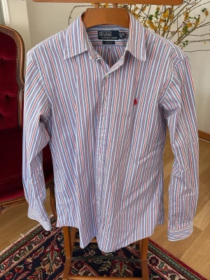Randig skjorta från Polo Ralph Lauren - Klassisk skjorta från Polo Ralph Lauren med blå, röda och vita ränder. Skjortan har lång ärm, krage och knäppning framtill. Broderad röd logga på bröstet. Tillverkad i bomull för en skön och luftig känsla.