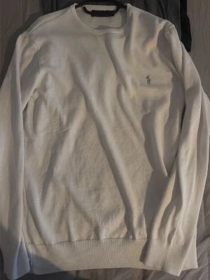 Vit tröja från Polo Ralph Lauren - Vit långärmad tröja från Polo Ralph Lauren med klassisk rund halsringning och broderad logotyp på bröstet. Tröjan är stickad och har ribbade muddar vid ärmslut och nederkant. Perfekt för dig som gillar stilrena basics.