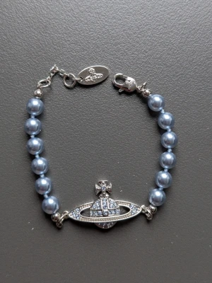 Blått pärlarmband Vivienne Westwood - Snyggt armband från Vivienne Westwood med blå pärlor och ikoniskt orb-hänge i silverfärg med gnistrande stenar. Låset är robust och har en liten tag med logotypen. Perfekt för dig som vill sticka ut med coola accessoarer.