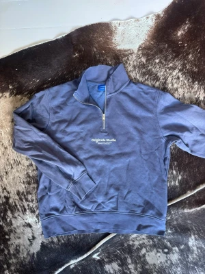 Blå halvzip hoodie från Jack & Jones - Snygg blå halvzip hoodie från Jack & Jones Originals Studio med vit logga på bröstet. Tröjan har hög krage, dragkedja framtill och ribbade muddar. Perfekt för en chill och avslappnad stil. Materialet känns mjukt och bekvämt mot huden.